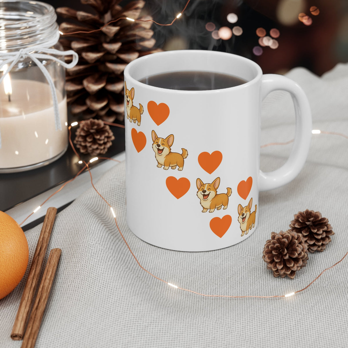 A white ceramic coffee mug printed with cartoon Corgi dogs and orange hearts, filled with black coffee and styled on a table with pinecones, a candle, cinnamon sticks, and fairy lights.