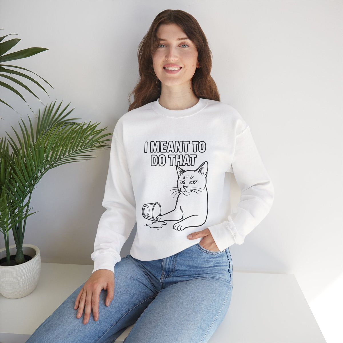 Woman wearing a white sweatshirt with a blue graphic and text, sitting on a white surface with a plant in the background.