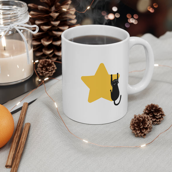 A white ceramic coffee mug with a yellow star and black cat graphic, filled with hot coffee and set on a tablecloth with pinecones, fairy lights, cinnamon sticks, and a candle in the background.