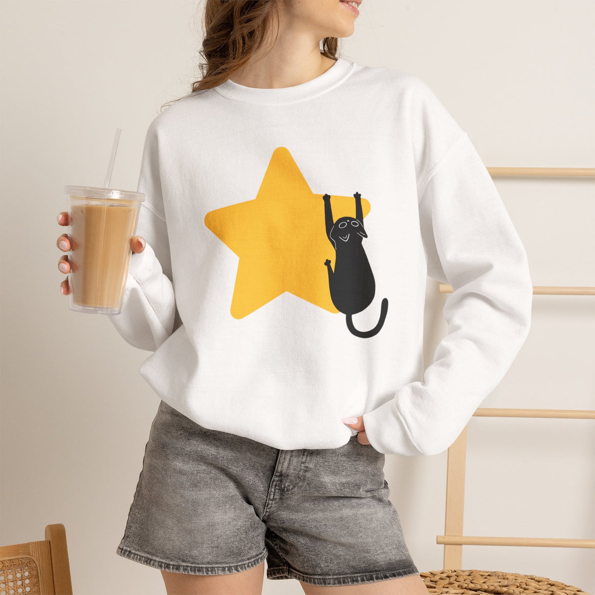 A woman wearing a white crewneck sweatshirt with a graphic of a black cat climbing a yellow star, holding an iced coffee in a cafe setting.