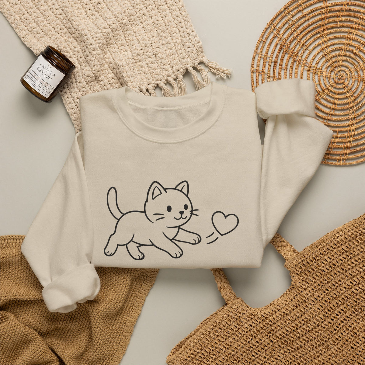 A folded cream sweatshirt  with a cat and heart design on the chest, styled in a flat lay with a woven blanket, a small candle jar, a woven mat, and a tan knit bag on a light neutral background.
