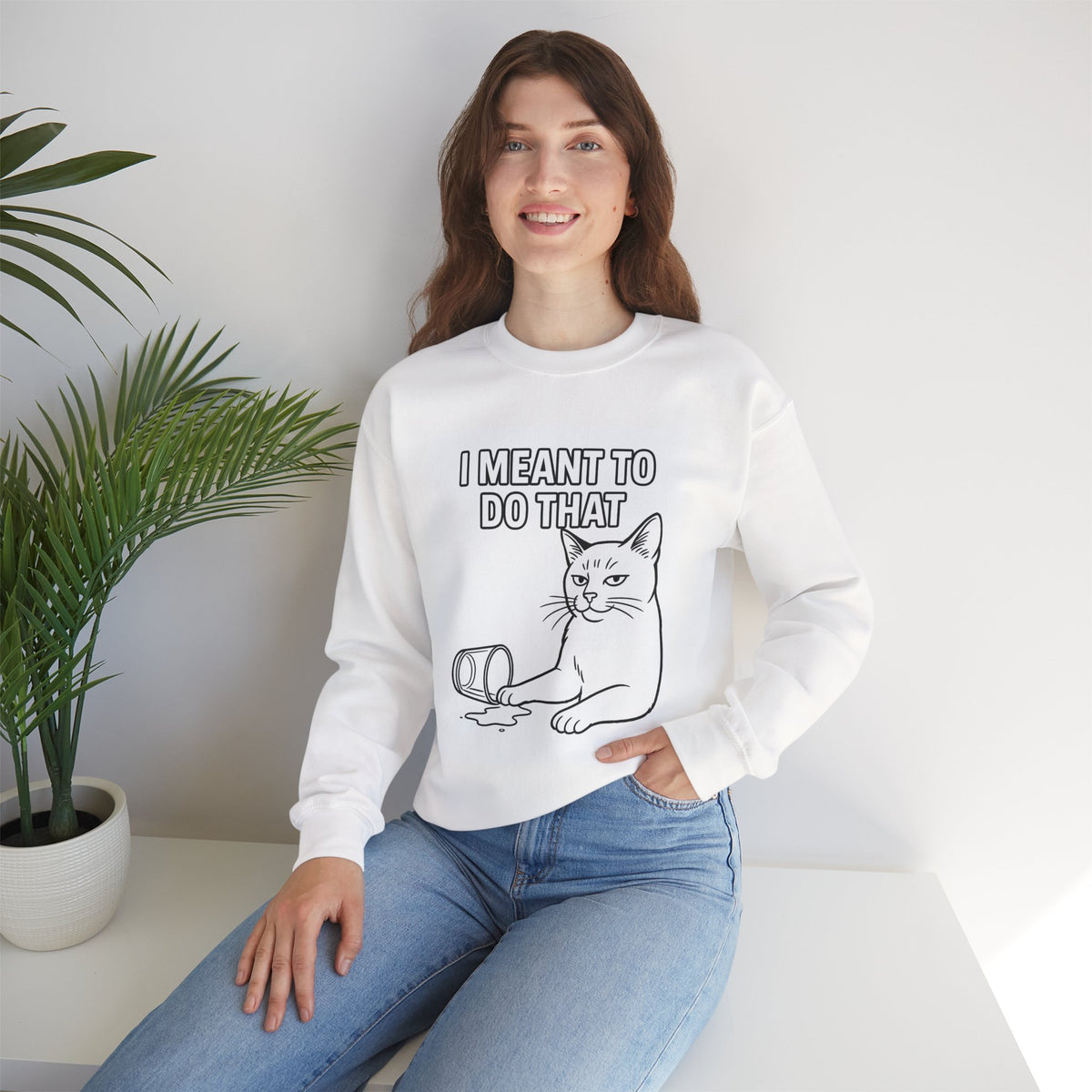 Woman wearing a white sweatshirt with a blue graphic and text, sitting on a white surface with a plant in the background.